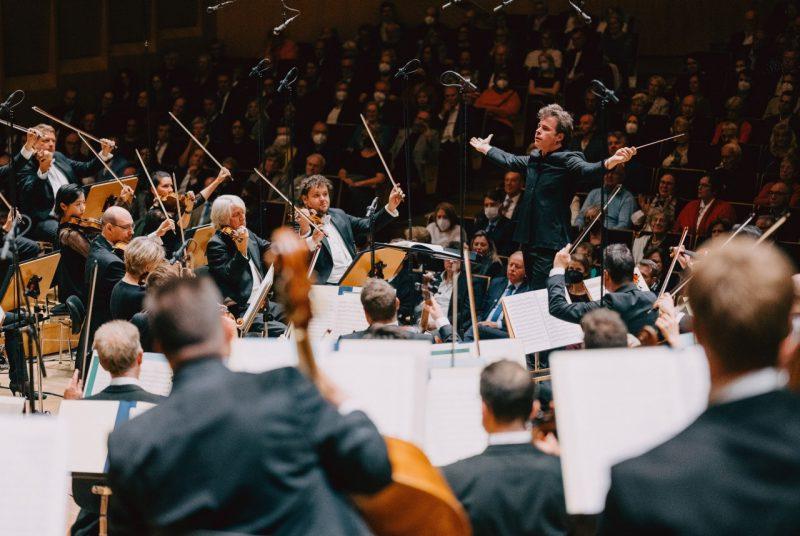 Conductor Jakub Hrůša, a middle aged white man with sandy brown hair, extends his arms out wide as he conducts the Bamberg Symphony during a live performance. All the string players watch him and their bows are raised in the air away from their instruments.