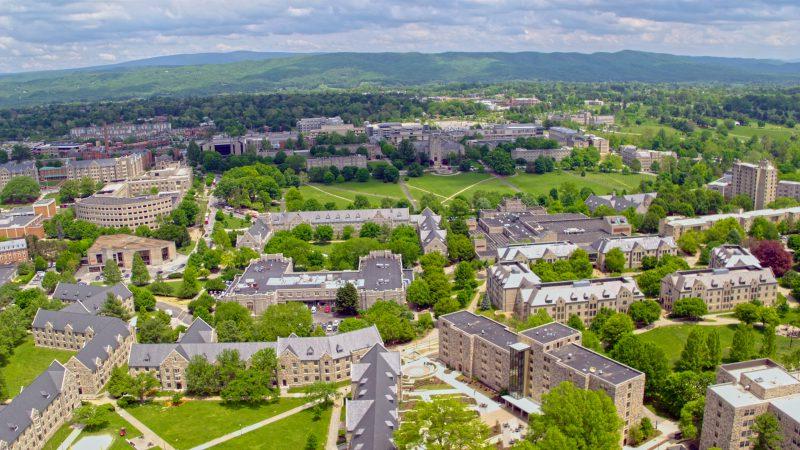 From an aerial view, gray Hokie Stone buildings are tucked between greenery in a mountain valley on a clear, sunny day.